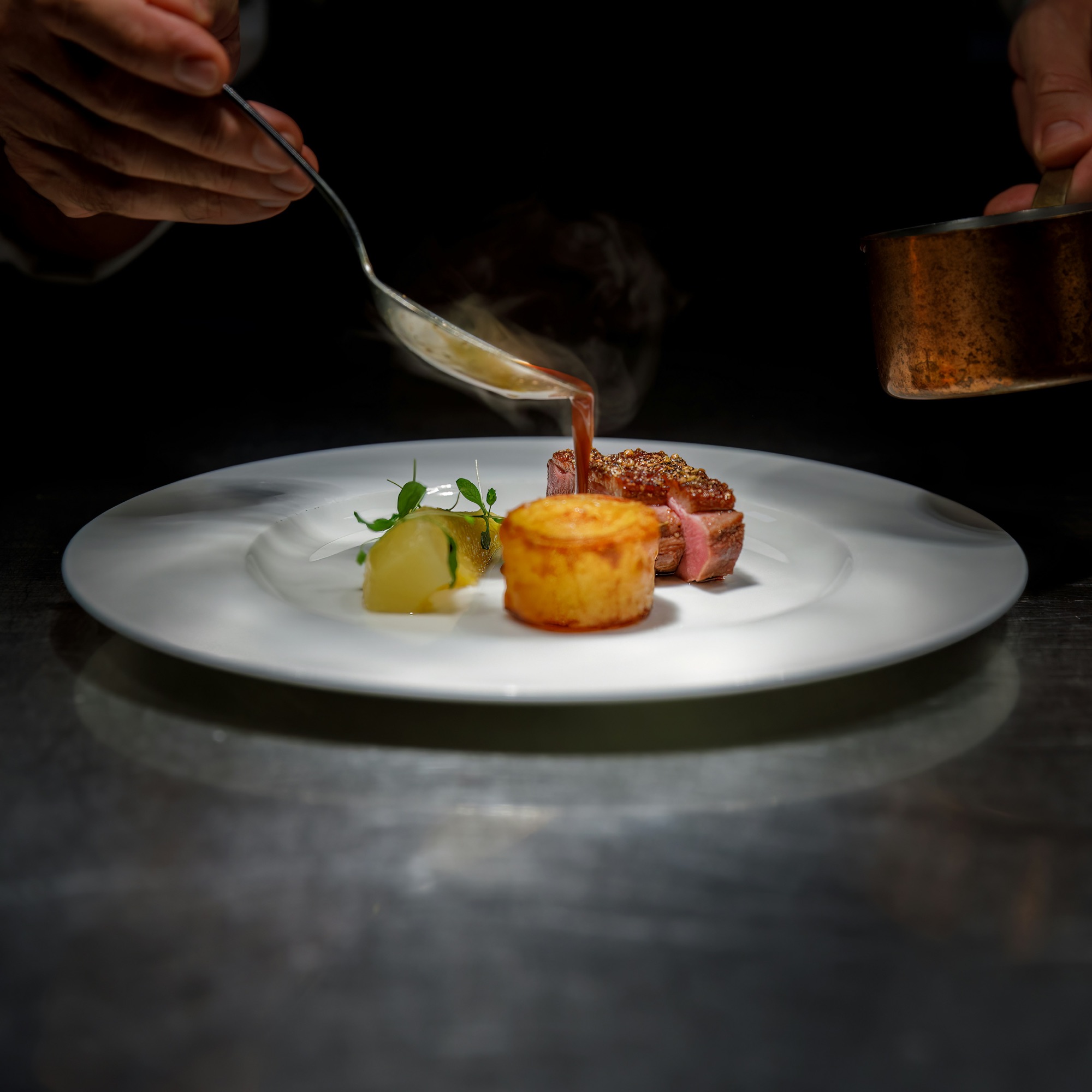A chef pours sauce over a plated dish in a dark dining room