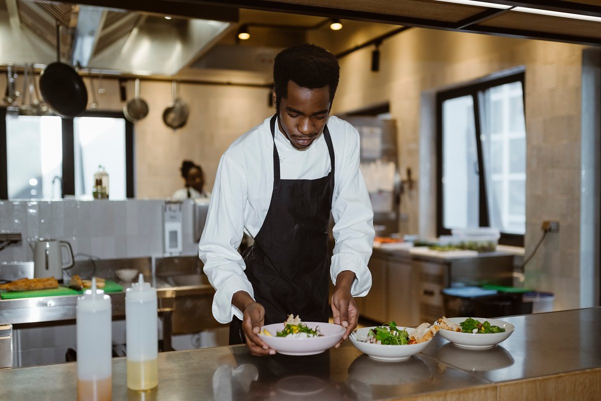 A young chef in a black apron sets down bowls of salad on a kitchen pass